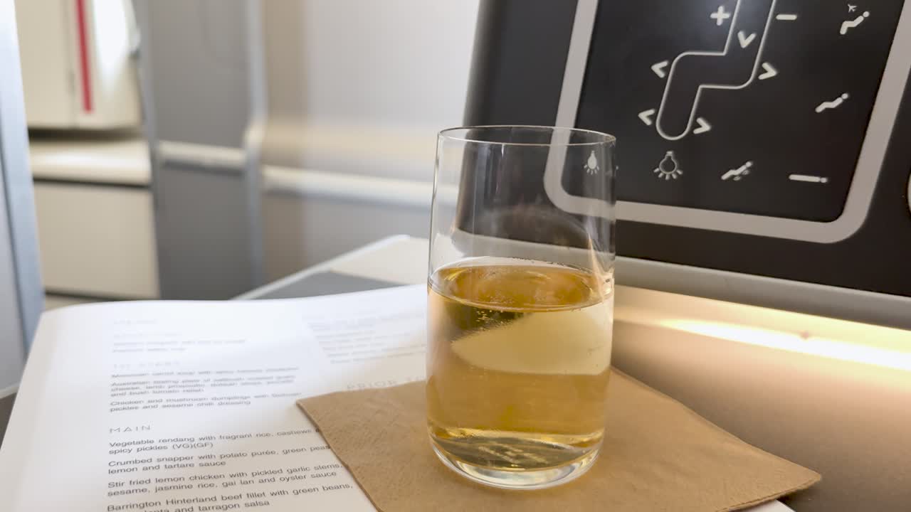 Glass of sparkling wine on napkin, airplane seat tray, soft daylight, minimal camera movement