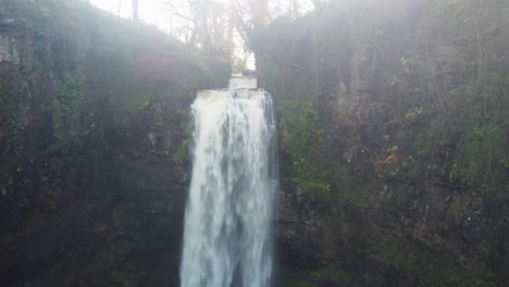 Cascada-Al-Amanecer-Con-Un-Dron-Aéreo-Que-Sube-Lentamente-Para-Revelar-Un-Río-Rápido-Y-Un-Sol-Dorado-En-Gales,-Reino-Unido-4k