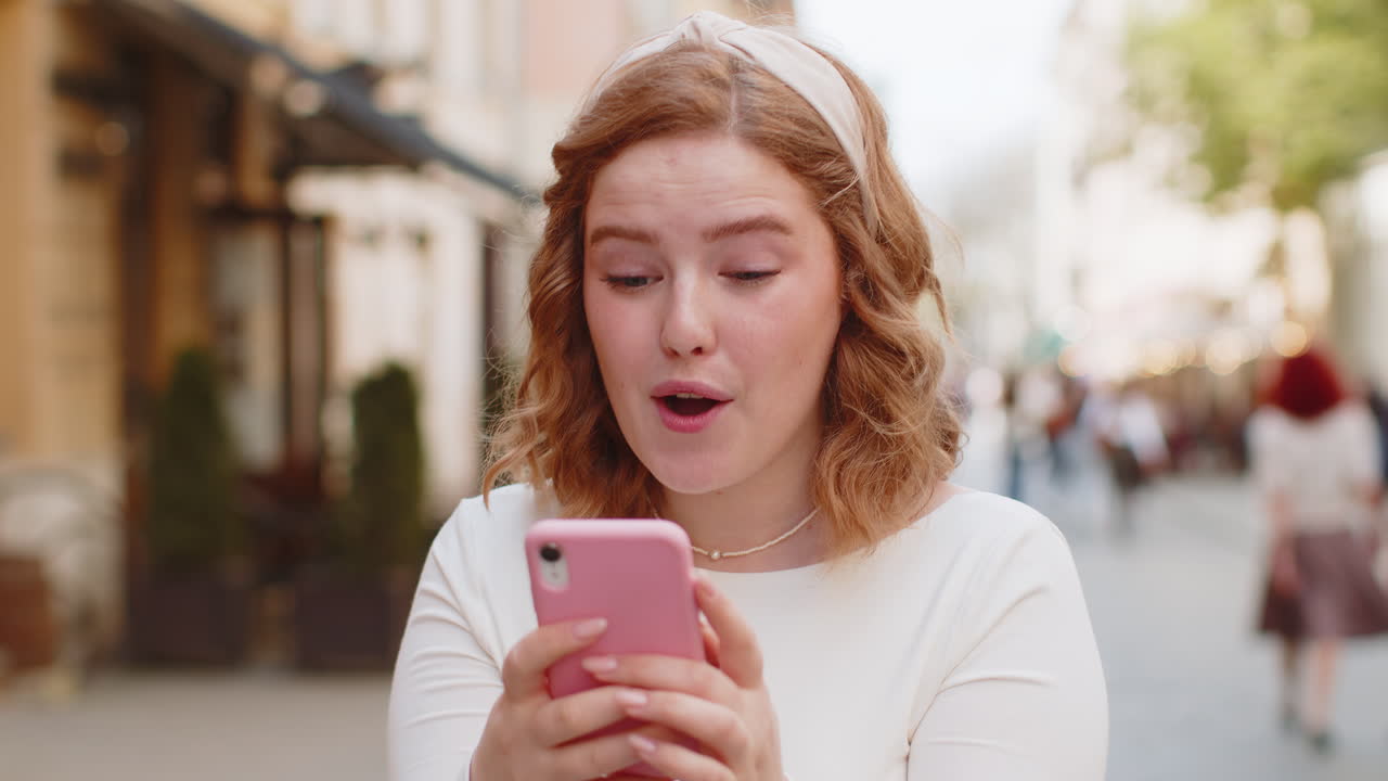 счастливая победительница взволнованная молодая женщина использует смартфон, празднуя победу хорошие новости на городской улице