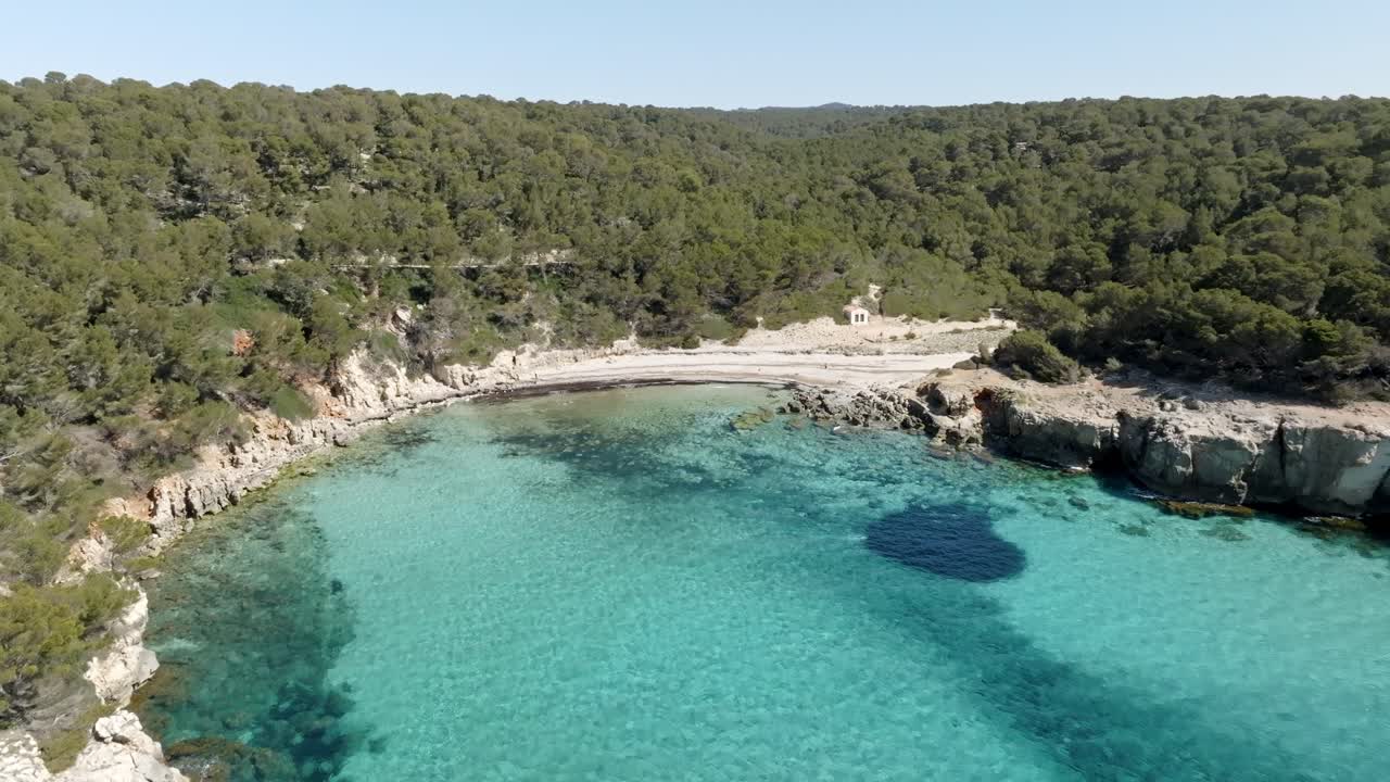 vista aérea de cala escorxada, la playa virgen en menorca, españa con agua verde y una colina cercana con árboles verdes y cielo claro azul en el fondo