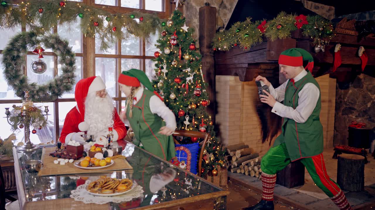 Santa Claus together with fairy elves. One elf is taking photo by retro camera. Female elf and Santa photographing on Christmas decorated room.