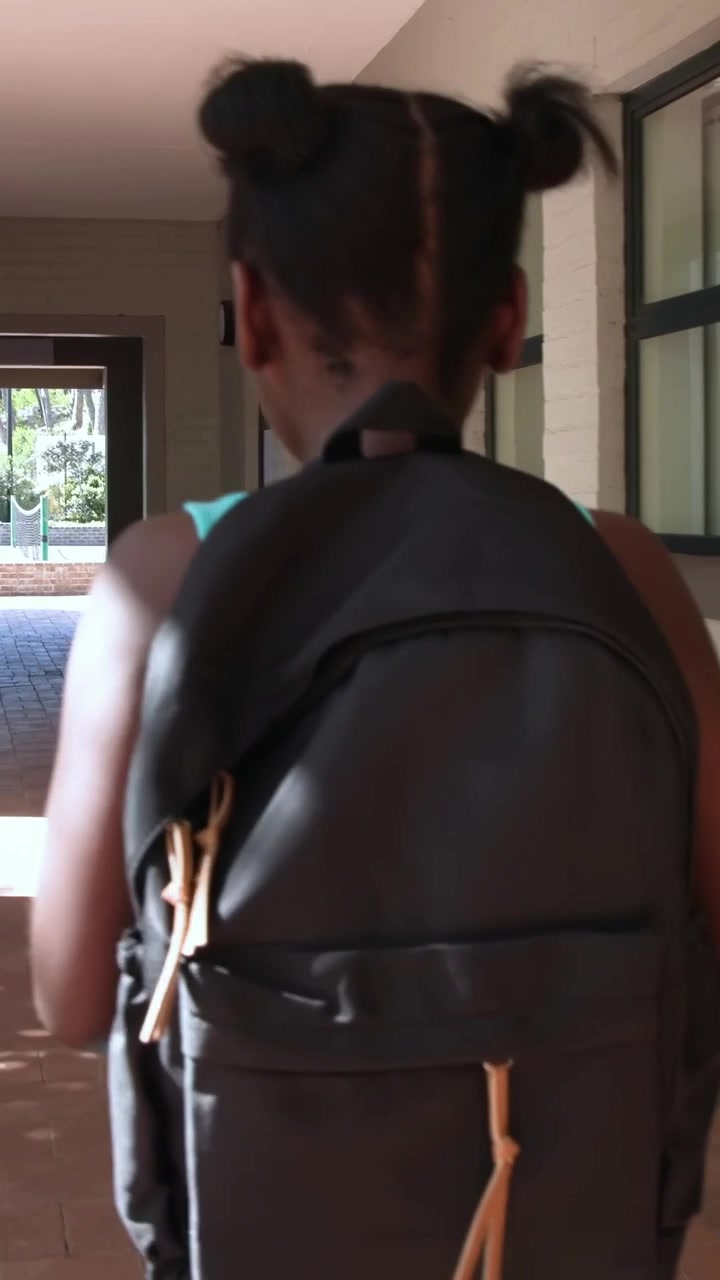 Vertical video: Walking girl in teal dress, sun drawing her toward hall exit, black pack tan tabs
