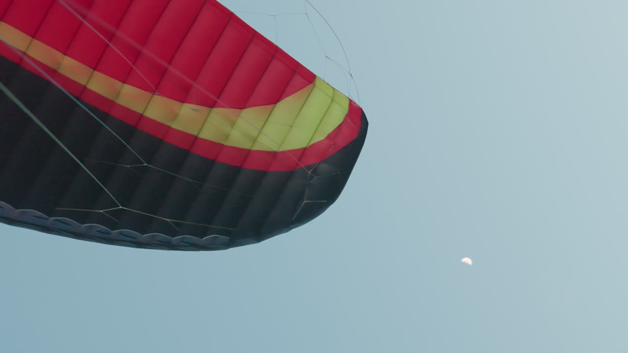 Colorful paraglider canopy graceful drifting across vast twilight sky with half moon overhead soft dusk light contrasting delicate rigging against serene cloud backdrop