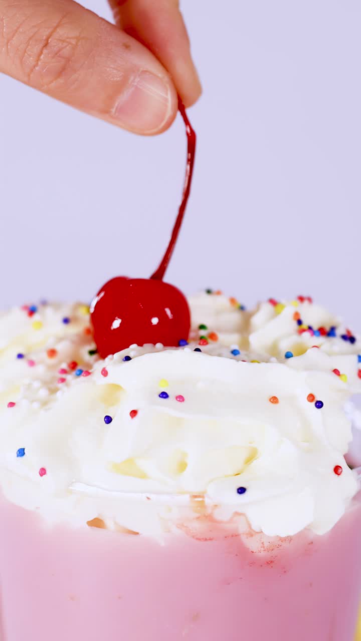 Primo piano verticale di una mano che guarnisce un milkshake rosa con panna montata, ciliegia e codette colorate.
