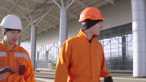 Two-construction-workers-in-orange-uniform-and-helmets-walking-through-the-construction-field-and-looking-over-plans-together.-Building-at-the-background