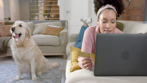 mujer biracial feliz con perro golden retriever usando portátil y tarjeta de crédito en casa, cámara lenta