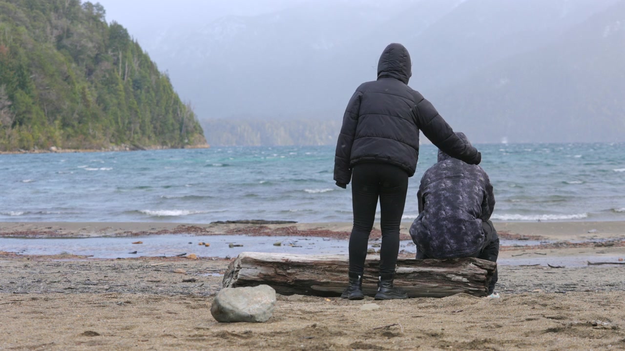Una persona conforta un'altra su una spiaggia fredda, entrambi di fronte all'oceano. La scena trasmette empatia e connessione. Ideale per narrazioni su supporto e amicizia.