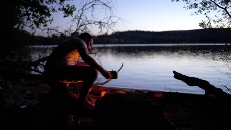 Einsamer-Camper-Hält-Einen-Topf-In-Der-Hand,-Während-Er-In-Der-Abenddämmerung-Auf-Dem-Campingplatz-Am-Seeufer-Feuer-Bläst