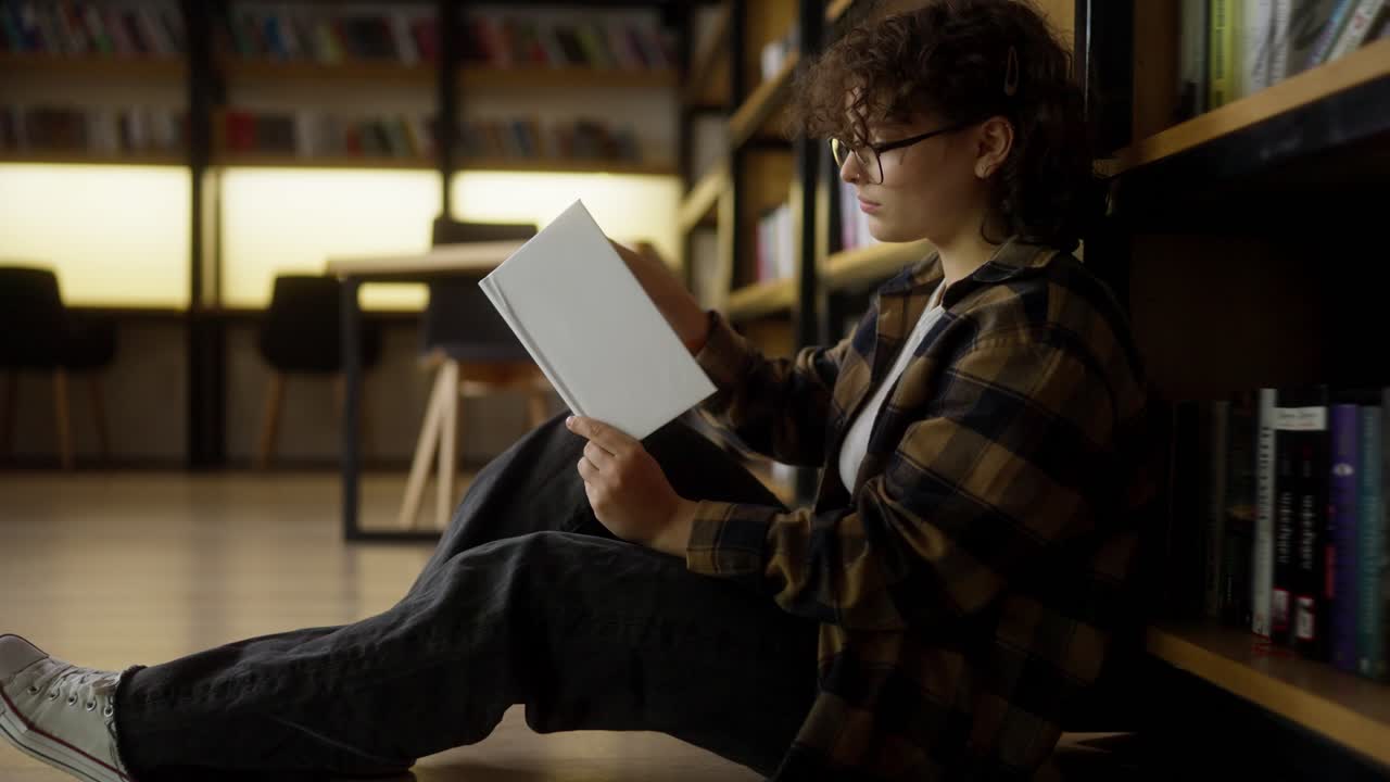 chica segura con cabello rizado y gafas se sienta cerca de los estantes con libros y lee uno en la biblioteca