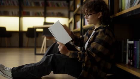 Confident-girl-with-curly-hair-and-glasses-sits-near-shelves-with-books-and-reads-one-in-the-library