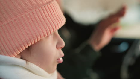 a close-up shot of a young girl wearing a pink beanie and jacket, seated inside a car. her expression is focused, with her lips slightly pursed as she looks ahead