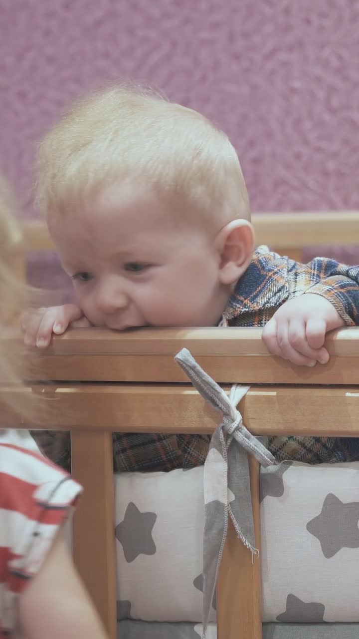 funny little girl jumps up at rocking cot playing with junior brother in light children room at home closeup
