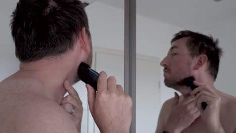 man shaving his beard in front of a mirror with an electrical razor