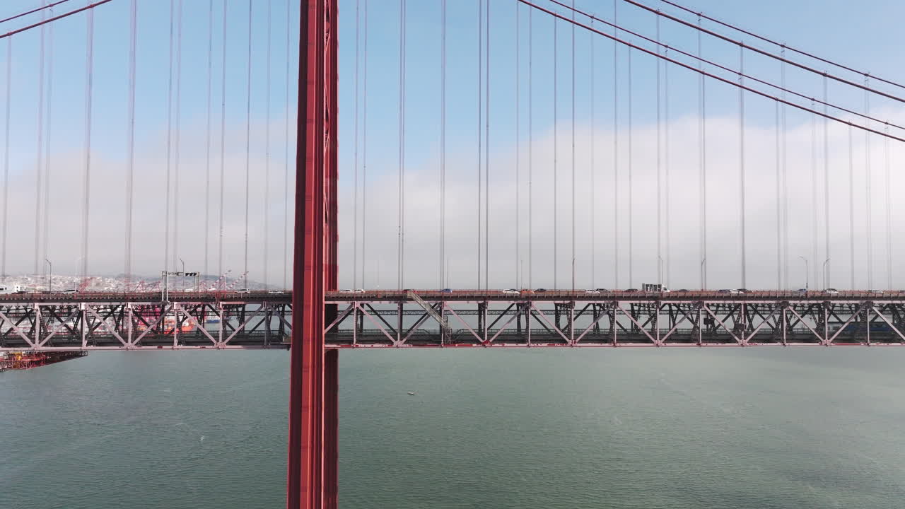 Aerial drone shot towards the 25th April Bridge in Lisbon, Portugal, Europe. Sunny and bright with clouds in the background. Cars driving on bridge, side view. Close-up perspective
