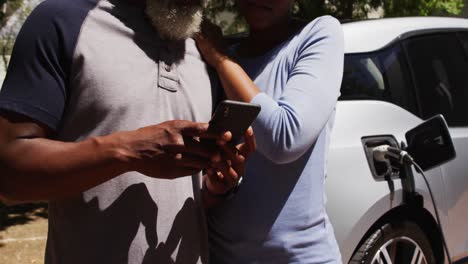sección media de una pareja afroamericana cargando un coche eléctrico y usando un teléfono inteligente en un día soleado