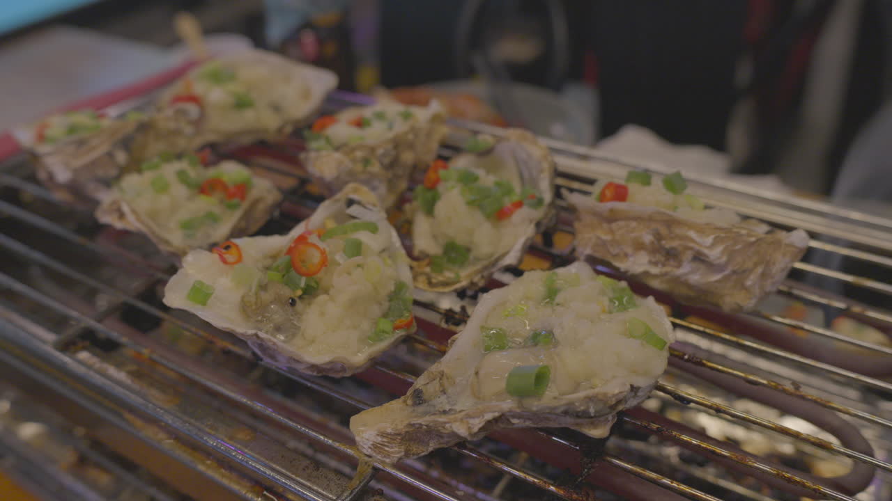 Sizzling grilled oysters topped with green onions and chili on metal rack, culinary close-up.