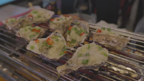 Sizzling grilled oysters topped with green onions and chili on metal rack, culinary close-up.