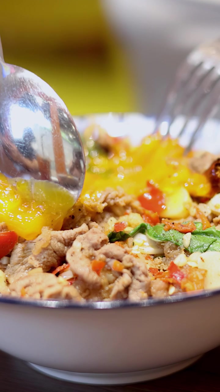 A spoon and fork break a runny egg yolk over a vibrant Thai rice bowl with shrimp, garlic, chili, and herbs in bright indoor lighting