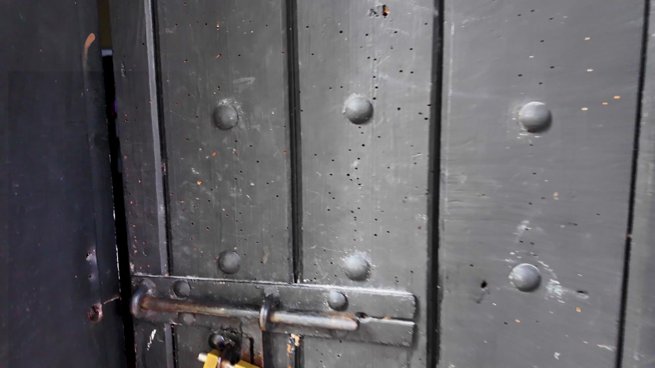 Approach to a rustic wooden door with a yellow padlock and hanging keys, slowly opening to reveal a dark, mysterious room within a traditional cottage.