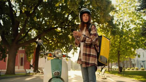 A-brunette-courier-girl-in-a-checkered-shirt-in-a-White-motorcycle-helmet-and-with-a-yellow-bag-stands-near-her-moped,-looks-at-the-phone-screen-and-around-to-understand-where-to-deliver-her-order