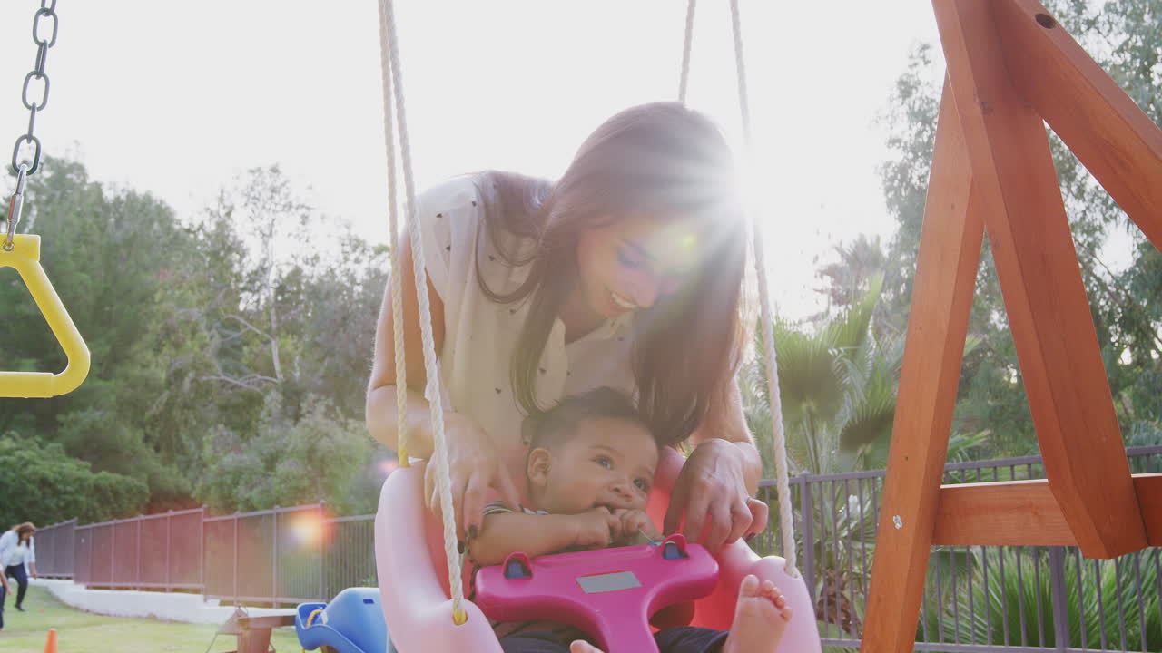 joven madre hispana empujando a su bebé en un columpio en un patio de recreo en el parque, de cerca