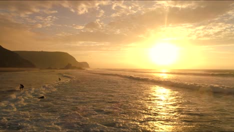 Surfer-Aus-Dem-Surfcamp-In-Neoprenanzügen-Kämpfen-Gegen-Die-Wellen-Und-Springen-Zur-Goldenen-Stunde-Praia-De-Cardoama-In-Sie-Hinein,-Während-Die-Wellen-Hereinbrechen