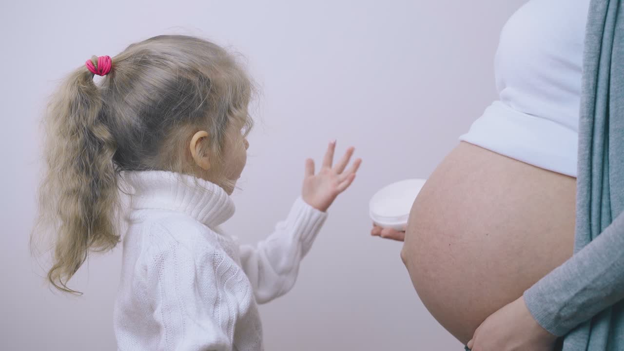 작은 소녀는 손가락으로 임신 한 엄마의 배에 로션을 적용합니다.