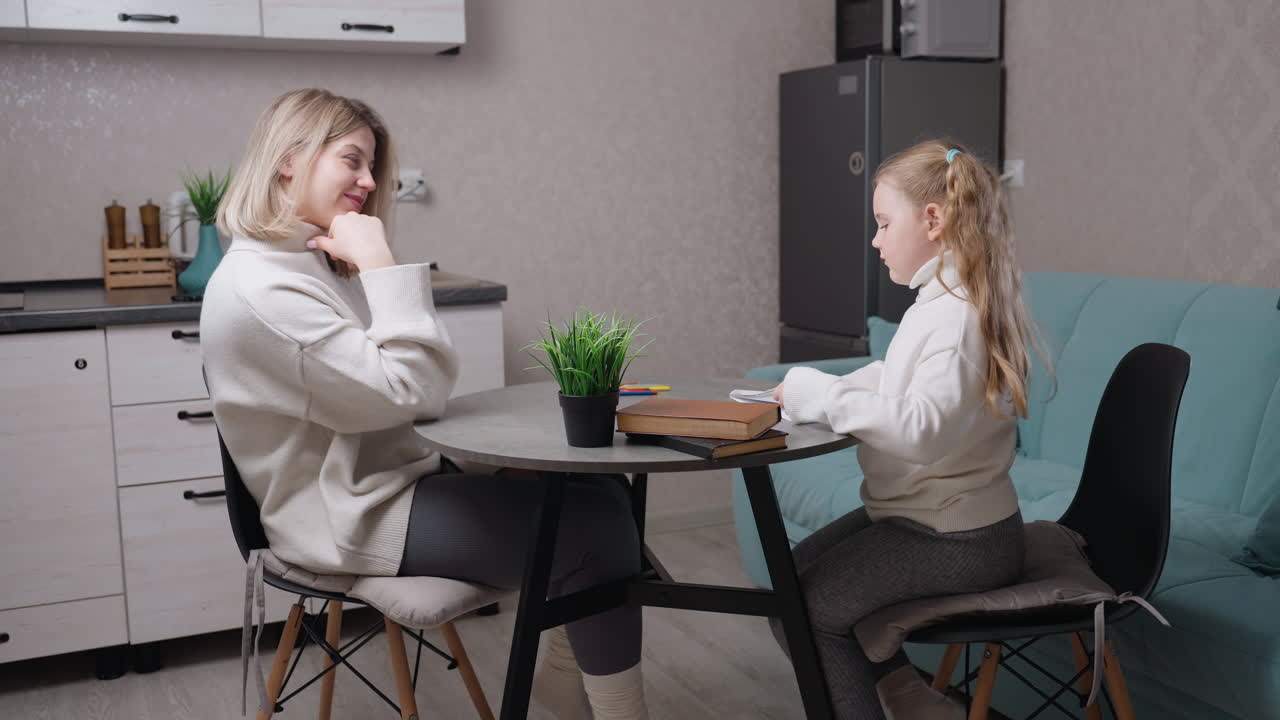 Mother and daughter in cozy kitchen sitting at round table, daughter flipping sketchbook pages while mother smiles warmly, family bonding moment with books, pencils, and small green plant on table