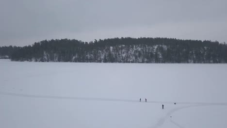 Toma-Aérea-Amplia-De-Cuatro-Amantes-De-La-Naturaleza-Disfrutando-De-Las-Temperaturas-Frías-Y-Nítidas-Durante-Un-Paseo-Matutino-En-Un-Vasto-Lago-Congelado