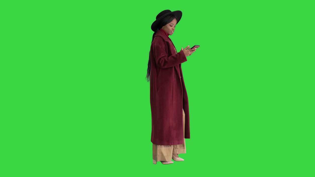 Linda jovem afro-americana estilosa com casaco e chapéu preto, digitando no celular enquanto caminha, em Tela Verde, Chroma Key.
