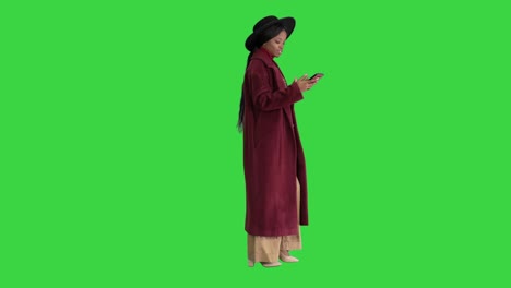 beautiful african american fashion girl in coat and black hat texting on her phone while walking on a green screen, chroma key