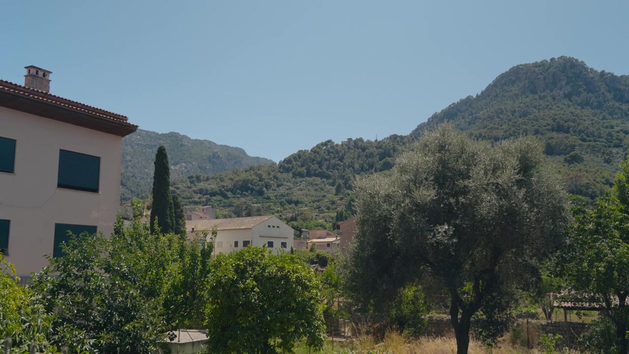 Ampia inquadratura su giardini e tetti verso le colline boscose sotto un cielo azzurro e limpido. Paesaggio rurale mediterraneo pacifico a mezzogiorno, ideale per sfondi di viaggio