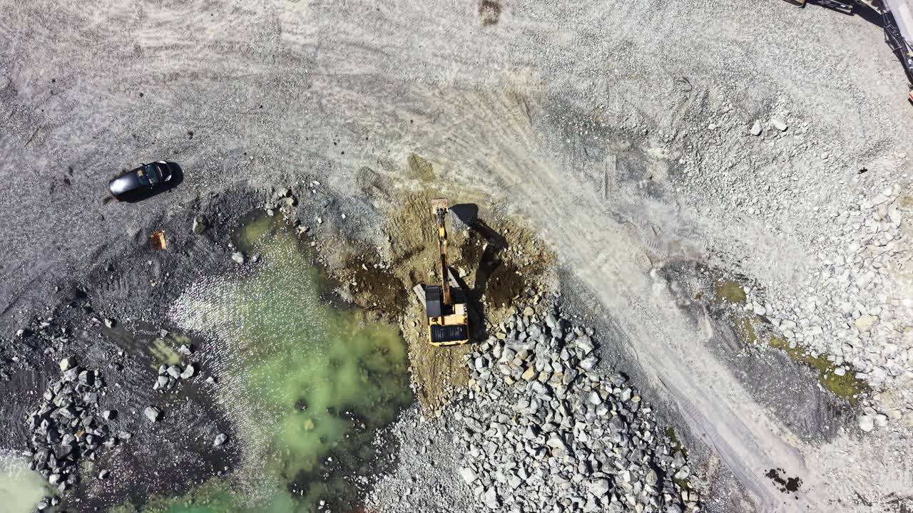 Workers extract granite at a quarry with machinery near a green water pond during daylight in a remote location