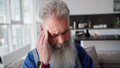 primer plano de un anciano con cabello gris y barba llena en una camisa azul masajea su templo durante un dolor de cabeza mientras está sentado en un sofá en un apartamento moderno