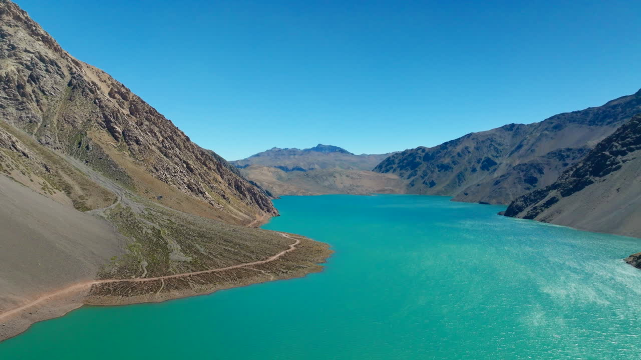 Drone flight over turquoise water of Embalse El Yeso reservoir in Cajon del Maipo, rugged Andes mountains and dirt road under clear blue sky, Chile. Aerial forward, copy space
