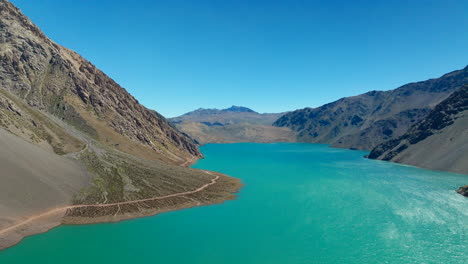 drone flight over turquoise water of embalse el yeso reservoir in cajon del maipo, rugged andes mountains and dirt road under clear blue sky, chile. aerial forward, copy space