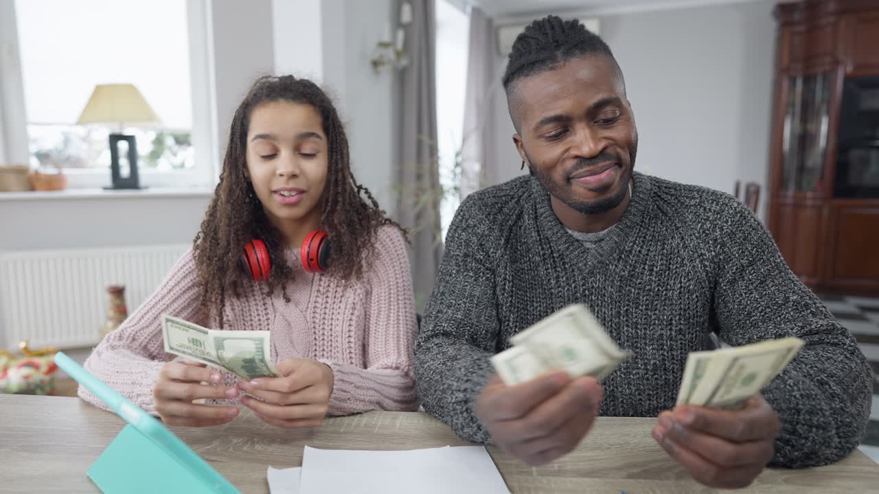 Front view portrait of African American rich man counting dollars sharing money with teen girl sitting at table in living room. Portrait of wealthy single father and teenage daughter at home with cash.