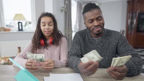 front view portrait of african american rich man counting dollars sharing money with teen girl sitting at table in living room. portrait of wealthy single father and teenage daughter at home with cash.