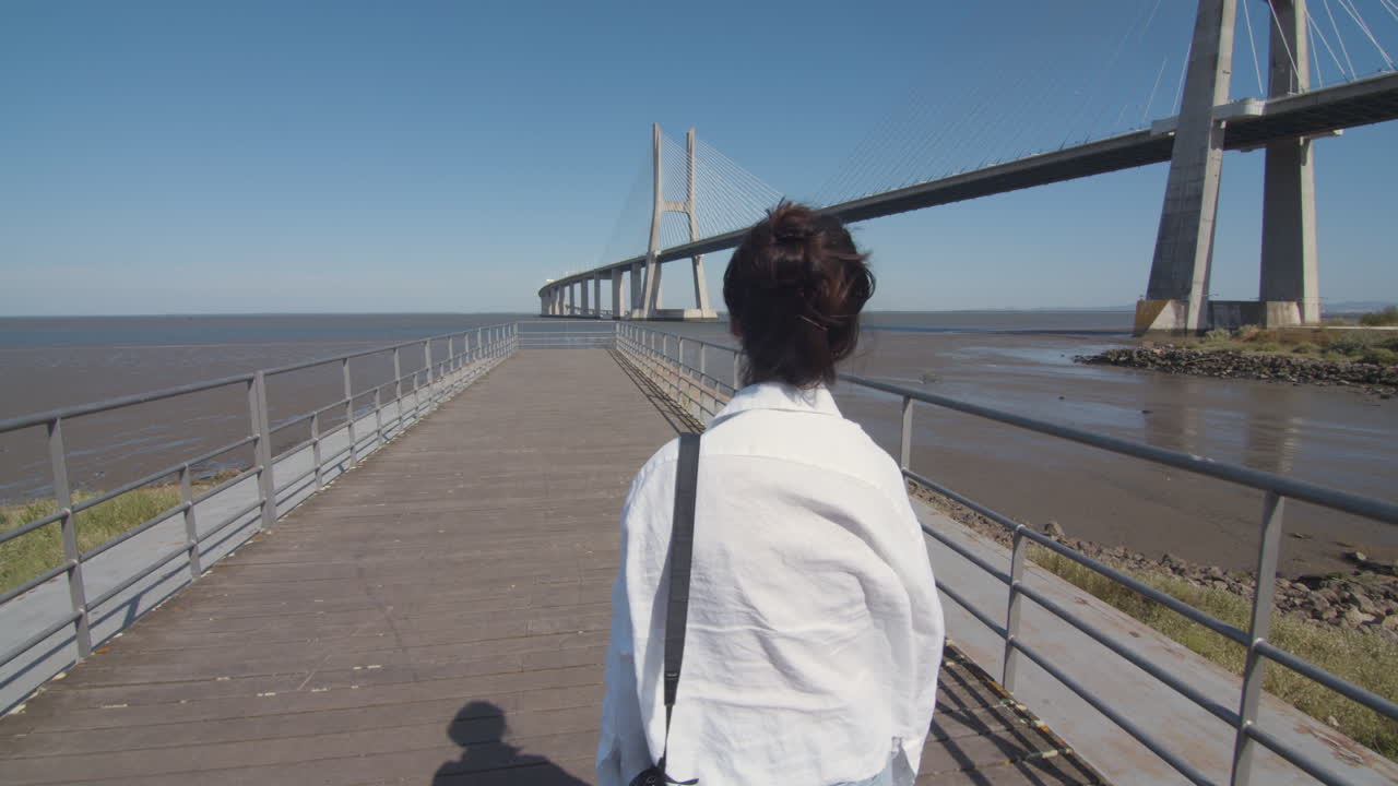 Follow shot of a young woman walking down the pier and enjoying the amazing view of the river with a bridge. Low tide.Tagus river. Contemplation in solitude. Architectural wonder