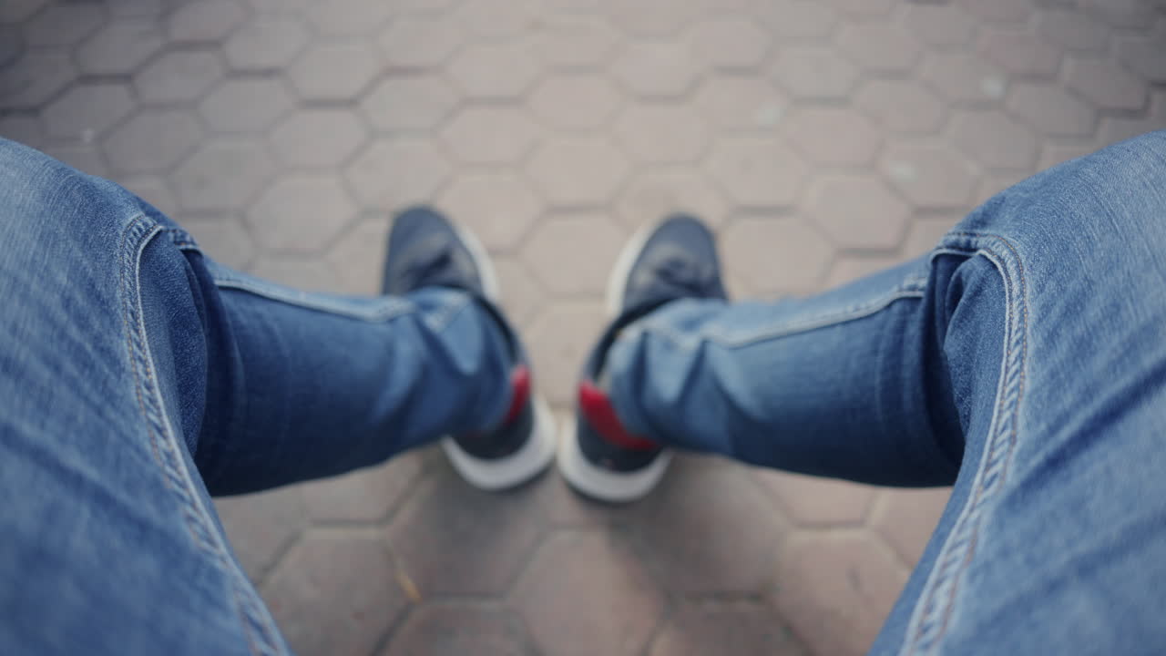 Vista superior de um homem sentado ao ar livre mostrando as pernas de jeans e tênis no pavimento.