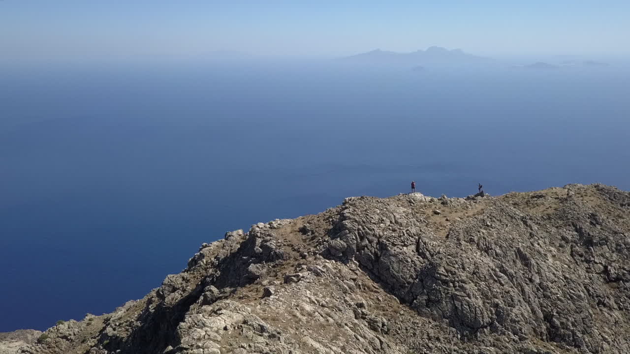 Aerial orbits hikers on mountain summit with view of boat on sea below