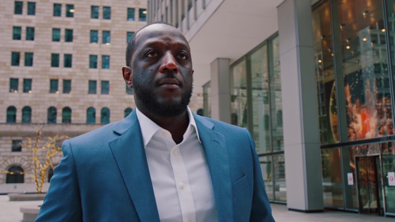 New York, USA, 21 May 2023: Black Businessman in Blue Suit Facing Camera in Urban Plaza. Man in a teal suit and white shirt faces the camera in a plaza with stone arched buildings and glass facade.