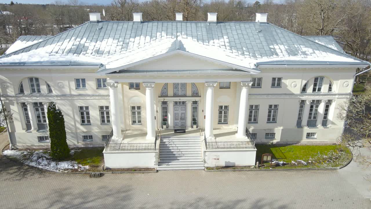 Aerial drone pullout footage of Saku manor or mansion during a sunny winter day. The white large historical arcitectural building has large white pillars and is in a green grass covered Estonian yard.