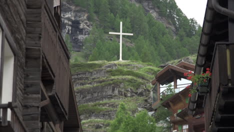 Tiro-Inclinado-Hacia-Arriba-De-Un-Pequeño-Pueblo-En-Zermatt-Y-Mostrando-Una-Gran-Cruz-Blanca-En-La-Cima-De-Una-Colina