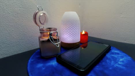 teléfono móvil negro que se enciende llamador entrante rodeado de luces y humidificador que cambia de colores y una vela encendida abierta en un frasco de vidrio