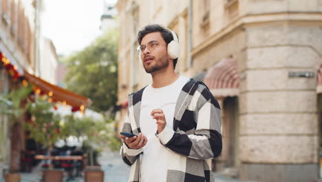 happy indian man putting on headphones choosing listening music in smartphone on city street