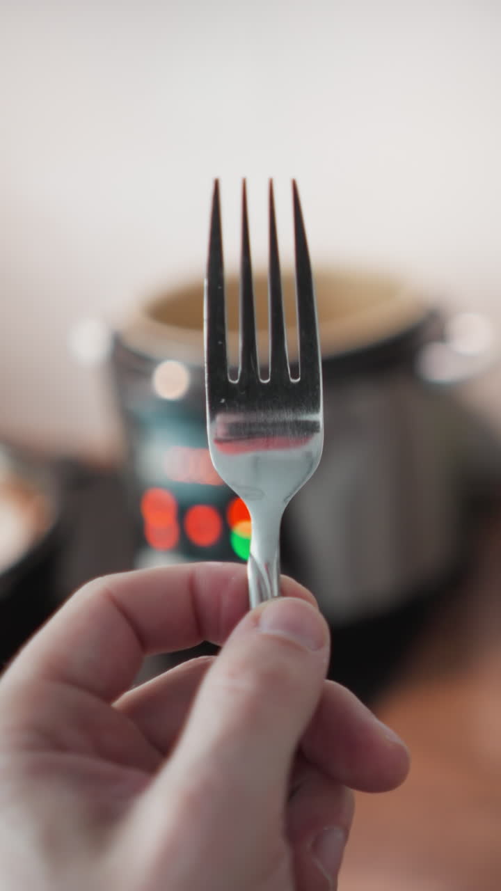 Hand-held POV video of a person twirling a fork with a multi-cooker's blinking lights in the blurred background