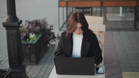 dama centrada en la computadora portátil al aire libre en un entorno urbano con su teléfono en la mesa, con un elegante abrigo negro, un pilar negro decorativo cerca, un recipiente de flores cerca y sillas en el fondo