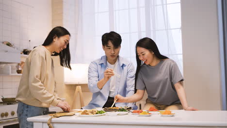 Three-Japanese-Friends-Sitting-Around-The-Kitchen-Counter-Cutting-And-Eating-Sushi-1