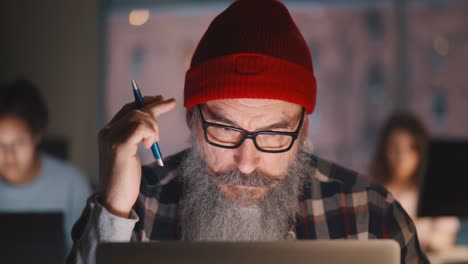 portrait of senior hipster bearded man holding pencil working on laptop in office